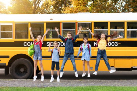 Téléchargez les photos : Groupe d'enfants heureux sautant devant le bus scolaire à l'extérieur, élèves joyeux avec des sacs à dos levant la main et regardant la caméra, garçons et filles joyeux s'amuser ensemble, pleine longueur avec espace de copie - en image libre de droit