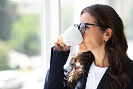 Photo for Closeup photo of brunette mature woman wearing eyeglasses and formal suit executive manager drinking coffee at office and looking through window, side view, copy space. Coffee break at work - Royalty Free Image