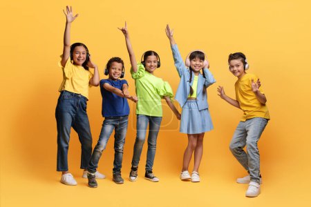 Photo for Happy carefree preteen diverse kids listening to music, dancing isolated on yellow background. Cheerful school aged multiethnic children boys and girls using wireless headphones, have party - Royalty Free Image