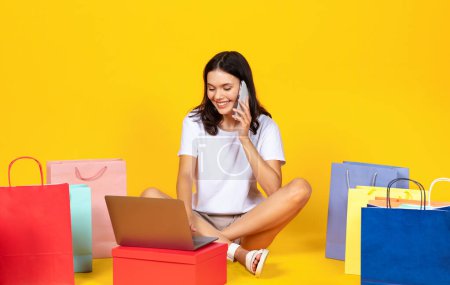 Photo for Happy young woman with shopping bags and laptop talking on cellphone, smiling shopaholic lady making online purchases, confirming web order, buying in internet, sitting on yellow studio background - Royalty Free Image