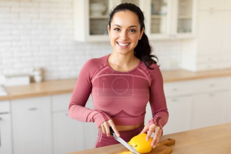 Photo for Happy young lady cutting fresh bell pepper on wooden board at kitchen table, preparing dinner from organic vegetables at home, smiling to camera while making healthy salad meal, wearing fitwear - Royalty Free Image