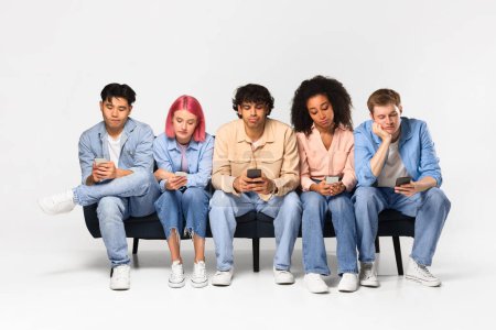 Photo for Sad friends looking at their cellphones, addicted multiracial people lost in gadgets, overuse social media networks, boring guys and ladies sitting on sofa over white background - Royalty Free Image
