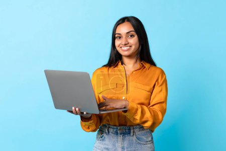 Foto de Publicidad de software informático. Mujer estudiante india alegre usando el ordenador portátil, posando con el gadget de la PC para el trabajo independiente y el E-aprendizaje, de pie en el fondo azul del estudio, sonriendo a la cámara - Imagen libre de derechos