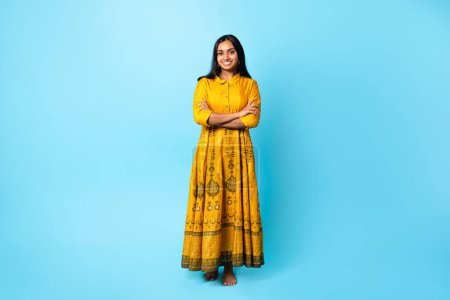 Foto de Atractiva mujer joven india posando en vestido amarillo tradicional con bordado, cruzando las manos y expresando confianza de pie sobre el fondo del estudio azul, sonriendo a la cámara. Longitud completa - Imagen libre de derechos