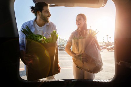 Foto de Joven pareja casada latina poniendo bolsas de papel llenas de comida saludable en el coche en el estacionamiento cerca del centro comercial o supermercado. Cónyuges cargando sus compras en coche - Imagen libre de derechos