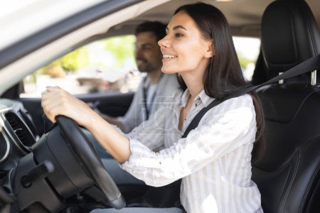 Aufgeregte junge brünette Frau sitzt am Autositz, schaut auf die Straße und lächelt, Fahrschülerin bereit für neue Lektion mit gutaussehendem Fahrlehrer, Seitenansicht