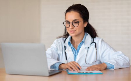 Photo for Young caucasian female nutritionist doctor in white coat making notes in clipboard, looking at laptop, sitting at table. Weight loss and health care, planning diet with professional - Royalty Free Image