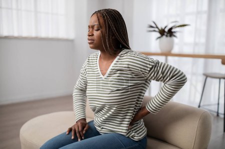 Photo for Unhappy upset young black woman with long braids sitting on couch at home, touching lower back, have strong pain, suffering from osteochondrosis, muscle strain, copy space - Royalty Free Image