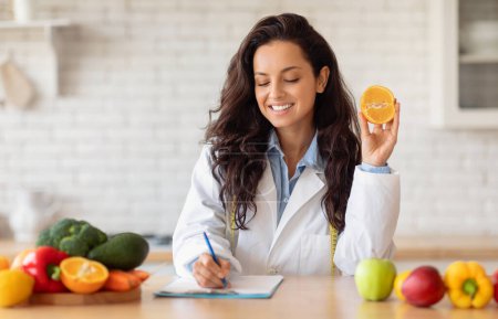 Photo for Cheerful european lady doctor nutritionist in white coat writing diet plan, recipe with organic fruits, holding orange. Health care, weight loss and professional recommendation - Royalty Free Image