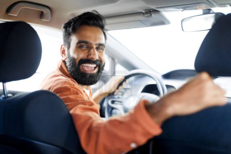Foto de Compra de coches. Alegre hombre árabe sonriendo en la cámara desde el asiento del conductor, disfrutando de su viaje sentado en el nuevo automóvil moderno en el interior. Propietario de automóviles conduciendo su vehículo con alegría, enfoque selectivo - Imagen libre de derechos