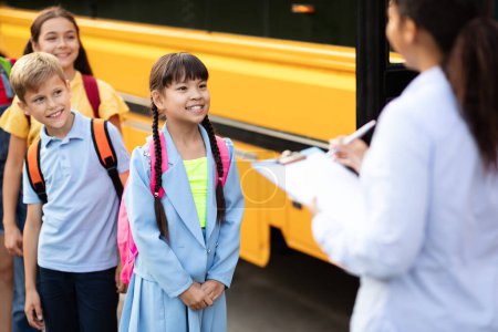 Téléchargez les photos : Une enseignante noire met à jour la liste de contrôle des enfants entrant dans un autobus scolaire, des écoliers heureux debout à leur tour près du véhicule, attendant de monter dans un autobus scolaire, se concentre sélectivement sur les élèves souriants - en image libre de droit
