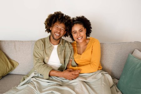Téléchargez les photos : Week-end romantique. Aimer les jeunes conjoints afro-américains partageant un câlin, couvert de couverture tout en étant assis sur le canapé, souriant à la caméra, posant à l'intérieur de la maison. Mariage heureux et relation d'amour - en image libre de droit