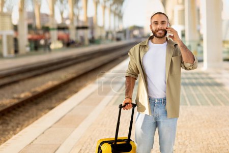 Photo for Smiling young caucasian guy with suitcase calls by phone, talk, waiting for transport on train station. Travel lifestyle, device for trip, communication, good news, business - Royalty Free Image