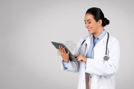 Téléchargez les photos : Gai médecin femme latine en uniforme blanc à l'aide d'une tablette numérique, femme médecin souriante avec gadget consulter les patients via un chat médical en ligne, posant sur fond gris, espace libre - en image libre de droit