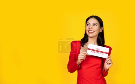 Téléchargez les photos : Heureuse jeune femme excitée tenant un certificat-cadeau et regardant à côté de l'espace de copie, belle femme joyeuse avec bon de réduction dans les mains posant isolé sur fond jaune studio - en image libre de droit