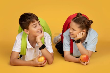 Téléchargez les photos : Élève adolescent européen positif avec sacs à dos et pommes, couché sur le sol à temps libre à l'école, isolé sur fond de studio orange. Etude, amitié et enfance, fruits et déjeuner - en image libre de droit