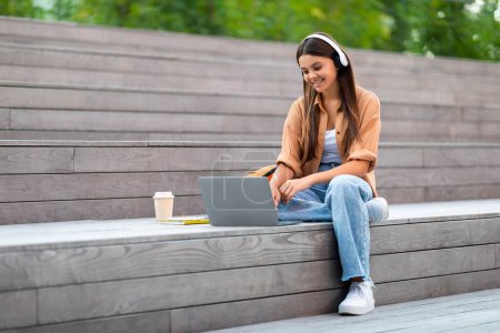 Téléchargez les photos : Positif assez élégant jeune femme hipster assis sur les escaliers de la zone urbaine de la ville, en utilisant ordinateur portable, casque sans fil à l'extérieur, boire du café à emporter, regarder webinaire, faire des devoirs, espace vide - en image libre de droit