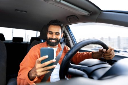 Foto de Alegre conductor árabe hombre mensajes de texto en el teléfono inteligente mientras conduce sentado en coche moderno en el interior. Hombre usando el teléfono móvil con la aplicación de navegador GPS navegando su nuevo automóvil, enfoque selectivo - Imagen libre de derechos