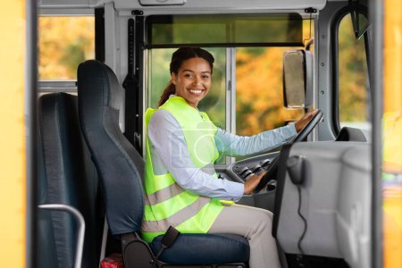Photo for Happy black female driver sitting inside of bus and looking at camera, smiling beautiful african american woman in reflecting clothing holding steering wheel, waiting passengers, enjoying her work - Royalty Free Image