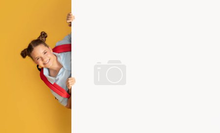 Téléchargez les photos : Heureuse écolière adolescente européenne regardant par derrière une grande bannière avec espace libre à l'école, isolée sur un fond de studio orange. Vente, recommandation pour étude, connaissance, annonce et offre - en image libre de droit