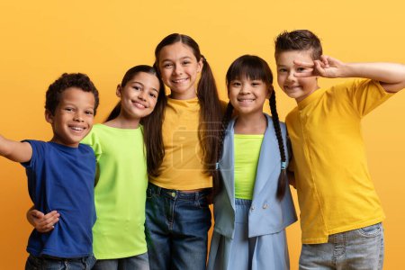 Happy childhood concept. Cute positive multiracial group of preteen kids taking selfie on yellow background. Cheerful school aged children boy and girls friends having together, smiling at camera