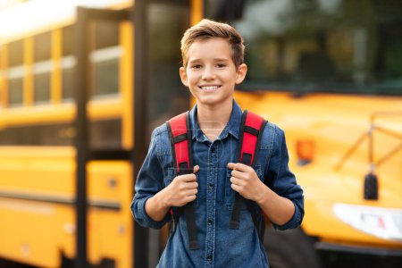 Foto de Feliz niño preadolescente sonriente de pie cerca de autobús escolar amarillo al aire libre, niño varón alegre con mochila posando afuera mientras va a clases, mirando a la cámara, colegial disfrutando del estudio, espacio para copiar - Imagen libre de derechos