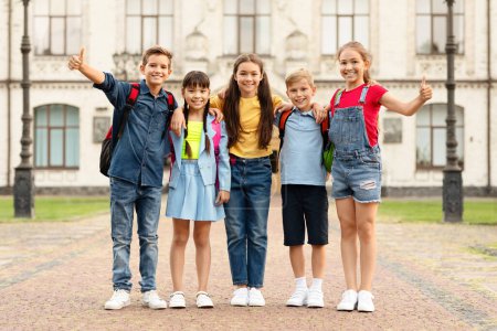 Photo for Group of happy multiethnic kids with backpacks posing outdoors, embracing and showing thumbs up, cheerful boys and girls standing against school building, enjoying study together, full length shot - Royalty Free Image