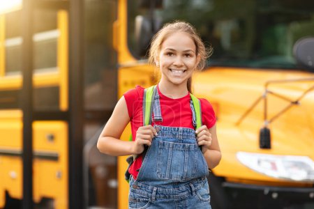 Foto de Sonriendo linda niña preadolescente de pie cerca de autobús escolar amarillo al aire libre, feliz niña con mochila posando afuera mientras va a clases, mirando a la cámara, colegiala disfrutando del estudio, espacio para copiar - Imagen libre de derechos