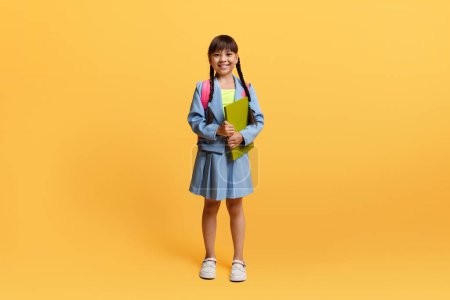 Téléchargez les photos : Joyeux pré-adolescente asiatique écolière avec sac à dos et exercisebook portant uniforme souriant isolé sur fond de studio jaune. Enfant chinois joyeux allant à l'école, copier l'espace - en image libre de droit