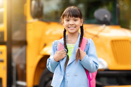 Foto de Linda chica asiática sonriente de pie cerca de autobús escolar amarillo al aire libre, niña feliz con mochila mirando a la cámara, posando afuera mientras va a clases, alumno alegre disfrutando de estudio, espacio de copia - Imagen libre de derechos