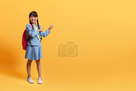 Téléchargez les photos : Jolie écolière coréenne preteen gaie en uniforme portant sac à dos pointant vers l'espace de copie pour la publicité ou le texte et souriant, isolé sur fond jaune. Éducation des enfants, concept scolaire - en image libre de droit