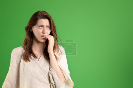 Photo for Worried confused young european lady in casual bites fingers, nervous, look at free space, isolated on green studio background. Stress, fear, expectation bad news, human emotions, ad and offer - Royalty Free Image