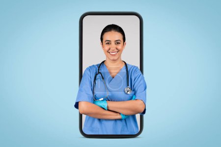 Foto de Servicios médicos modernos. Feliz joven doctora latina en uniforme azul con estetoscopio sonriendo a la cámara, posando con los brazos cruzados en la pantalla grande del teléfono celular, pancarta - Imagen libre de derechos