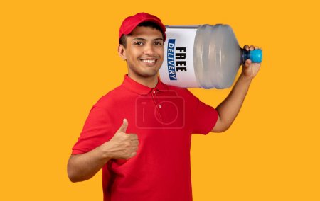 Téléchargez les photos : Livraison d'eau. Joyeux coursier hispanique tenant une grosse bouteille d'eau sur l'épaule, les pouces levés, coiffé d'une casquette rouge et d'un uniforme posant sur fond jaune. Livreur livre et approuve l'eau - en image libre de droit