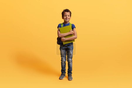 Téléchargez les photos : Préadolescent mignon enfant afro-américain allant à l'école, portant un sac à dos, tenant des cahiers d'exercices, isolé sur fond jaune, espace de copie. Éducation des enfants, école - en image libre de droit