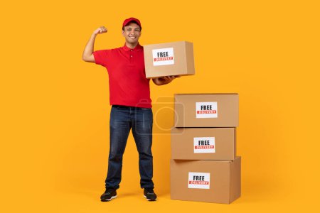Photo for Free Delivery Offer. Cheerful Middle Eastern Deliveryman Holding Carton Box Showing Biceps Muscles On Arm, Standing Near Stack Of Cardboard Boxes On Yellow Studio Background, Smiling At Camera - Royalty Free Image
