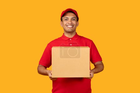 Téléchargez les photos : Mexicain livraison homme en uniforme rouge tenant boîte en carton, donnant colis souriant à la caméra, la livraison de commande en ligne au client sur fond jaune en studio. Expédition rapide et service de livraison - en image libre de droit