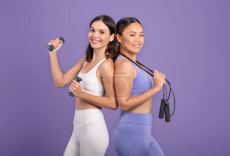 Foto de Jóvenes mujeres asiáticas y caucásicas en ropa deportiva de pie espalda con espalda con campanas y saltar la cuerda, mirando y sonriendo a la cámara, posando sobre fondo púrpura - Imagen libre de derechos