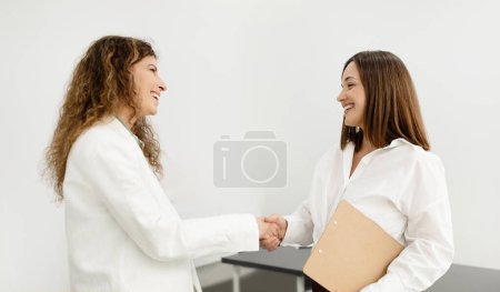 Photo for Business Deal. Two Smiling Businesswomen Meeting And Shaking Hands In Agreement, During Job Interview, Standing At Modern Office Interior, Wearing White Formal Wear. Partnership Concept - Royalty Free Image