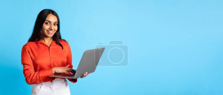 Foto de Freelance, Oferta Online. Feliz señora india que sostiene el ordenador portátil que navega por Internet y sonríe a la cámara, trabajando y estudiando a través de la computadora, posando con PC en el fondo del estudio azul. Panorama, Espacio vacío - Imagen libre de derechos