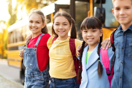 Foto de Grupo de niños felices multiétnicos de pie al aire libre cerca del autobús escolar, retrato de colegialas sonrientes y colegialas con mochilas abrazando y mirando la cámara, disfrutando del tiempo juntos, primer plano - Imagen libre de derechos