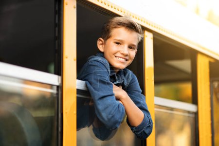 Foto de Guapo preadolescente mirando por la ventana del autobús escolar, alegre alumno masculino sonriendo a la cámara mientras se asoma fuera del autobús escolar amarillo, colegial listo para el viaje a casa después de las lecciones, primer plano - Imagen libre de derechos