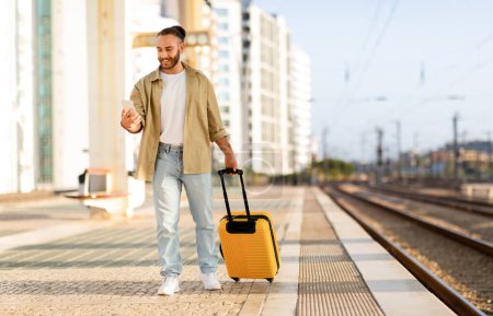 Photo for Cheerful young caucasian man with suitcase uses phone, chatting, waiting for transport on train station, full length. Work, travel lifestyle, trip with device app, summer vacation - Royalty Free Image