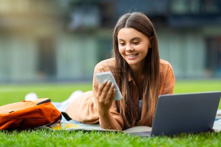 Téléchargez les photos : Cool joyeux jolie jeune étudiante utilisant des gadgets au parc, couché sur la pelouse avec un ordinateur portable, vérifier son smartphone, à la recherche de beaux cours éducatifs en ligne, défilement, espace vide - en image libre de droit