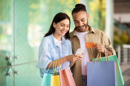 Foto de Sonriente joven europeo hombre y mujer con bolsas muestran tarjeta de crédito, usar el teléfono, disfrutar de compras y tiempo libre en el centro comercial. Recomendación financiera, venta y adictos a las compras, descuento y aplicación - Imagen libre de derechos