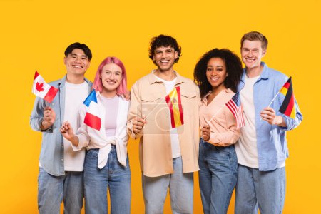 International friendship. Happy multiracial people holding different countries flags and smiling at camera, standing on yellow studio background. Modern education of students or immigrants