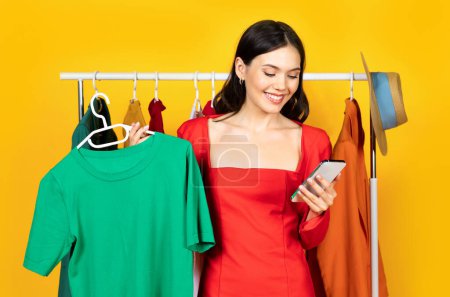 Photo for Smiling young woman using online shopping app on smartphone and holding hanger with t-shirt, happy female choosing new clothes in internet, standing near clothing rail over yellow studio background - Royalty Free Image