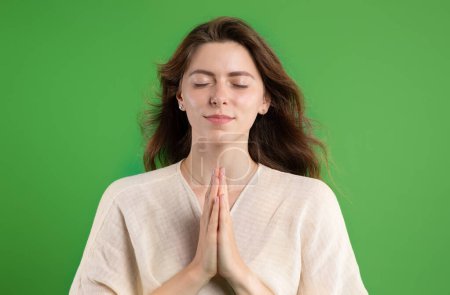 Foto de Jovencita europea positiva con los ojos cerrados rezando con el gesto de las manos, aislada en el fondo verde del estudio, de cerca. Pide deseos, peticiones y fe, anuncios y ofertas, emociones humanas - Imagen libre de derechos