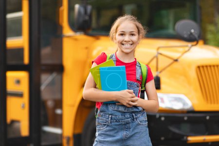 Téléchargez les photos : Retour à l'école. Joyeux sourire fille préadolescente debout près de l'autobus scolaire avec sac à dos et cahiers dans les mains, mignon enfant femelle posant à l'extérieur tout en allant à des cours, élève joyeux profiter de l'étude - en image libre de droit