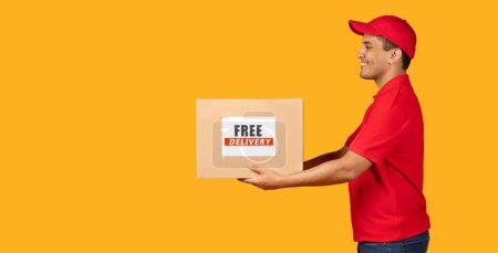 Photo for Free Delivery Offer. Hispanic Courier Guy Giving Carton Box Aside To Copy Space, Holding Carton Parcel On Yellow Studio Background. Side View Shot Of Deliveryman Shipping Purchase. Panorama - Royalty Free Image